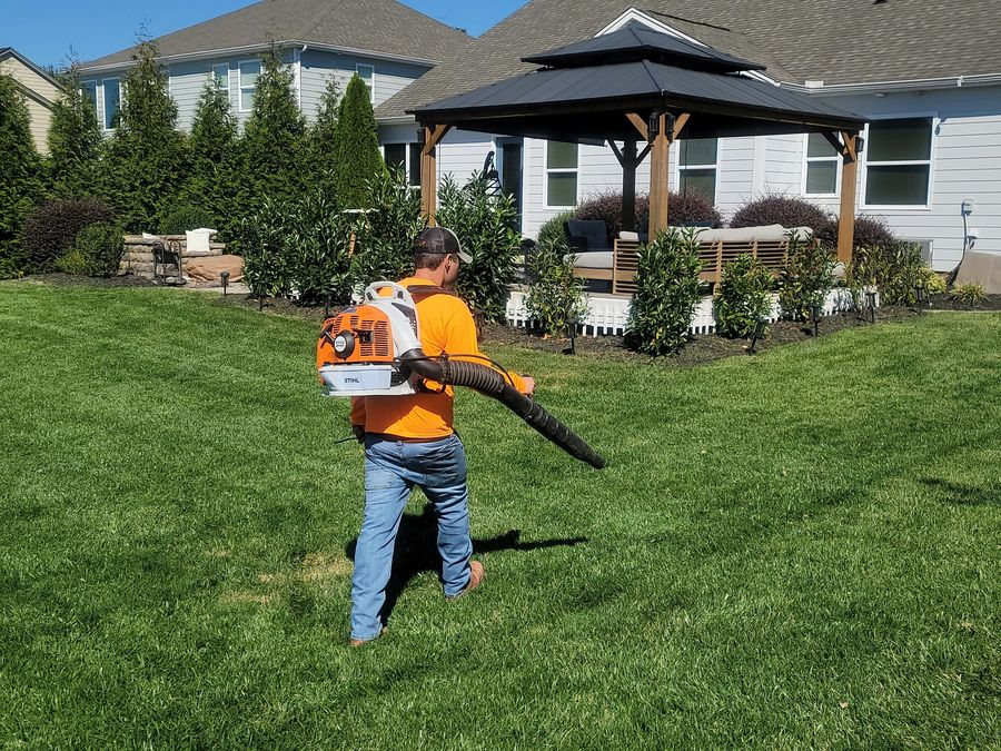 Crew member clearing clippings from a lawn beside planted beds and a covered patio